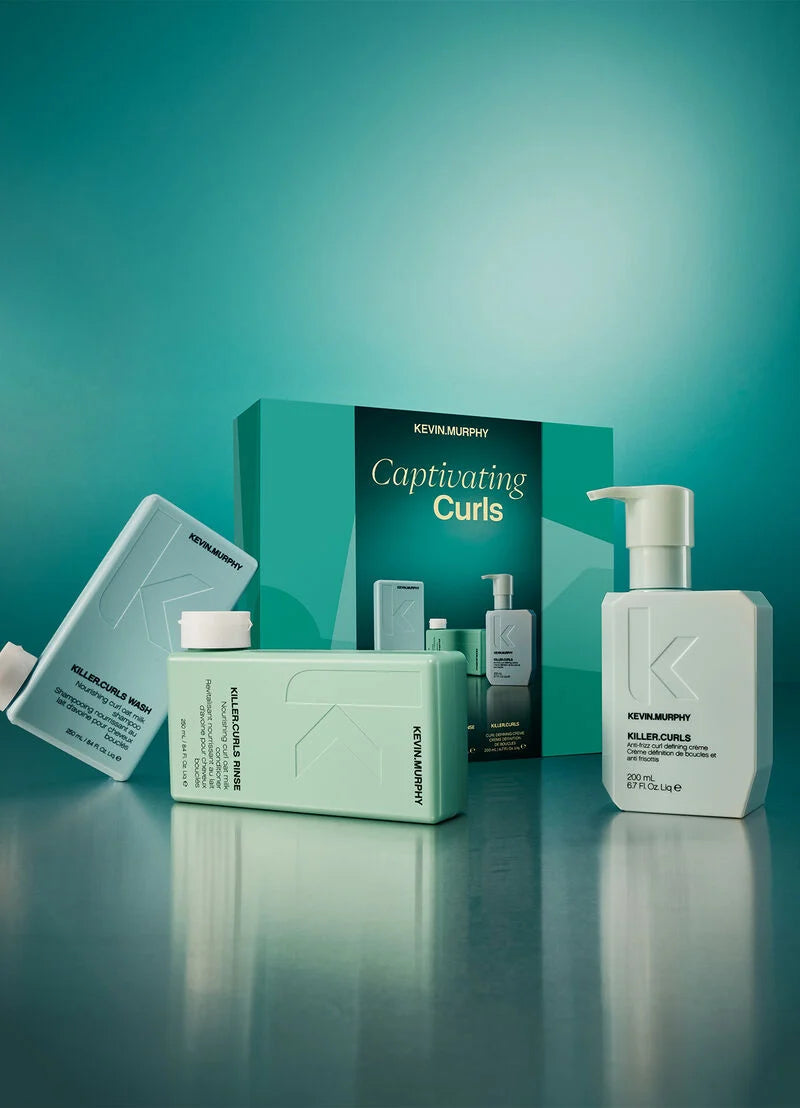 Kevin Murphy Captivating Curls haircare set featuring a teal and mint green box, a large white pump bottle, a mint green tub, and a white flat bottle, arranged on a reflective surface against a teal gradient background.