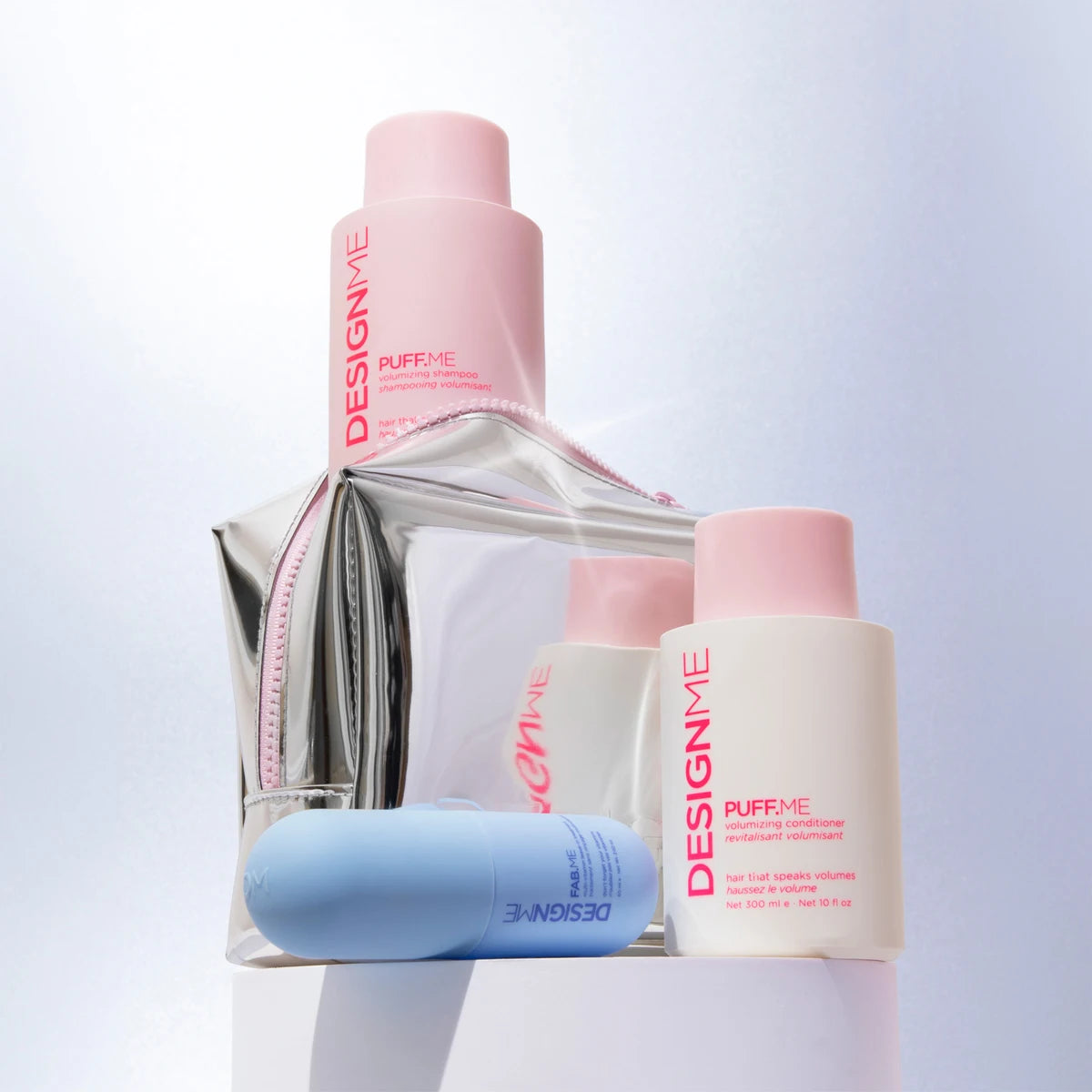 A collection of DesignMe Puff Me skincare products arranged on a white pedestal, including a tall pink bottle, a white pump bottle, a silver pouch, and a light blue tube, against a clean white background.