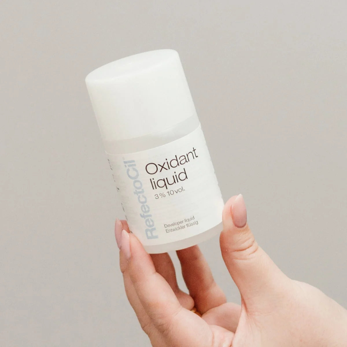 A hand holding a white bottle of ReflectoOil Oxidant liquid 3% 10 vol against a neutral light gray background.