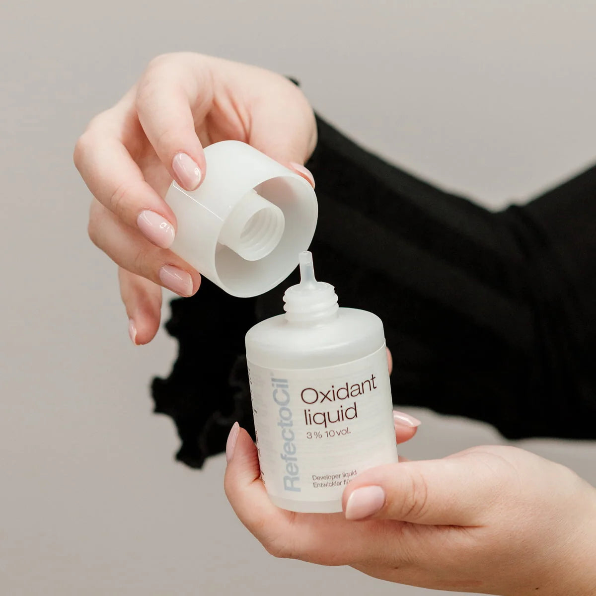 A close-up of a person holding a white bottle labeled 'Oxidant liquid' and unscrewing its cap to pour the liquid, with the brand name 'RefectoOil' visible on the bottle.
