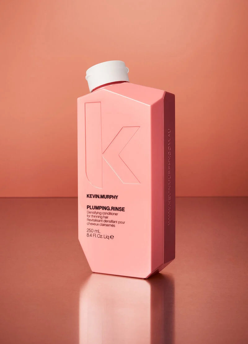 A pink Kevin Murphy Plumping Rinse bottle with a white cap, standing on a reflective surface against a solid peach-pink background.