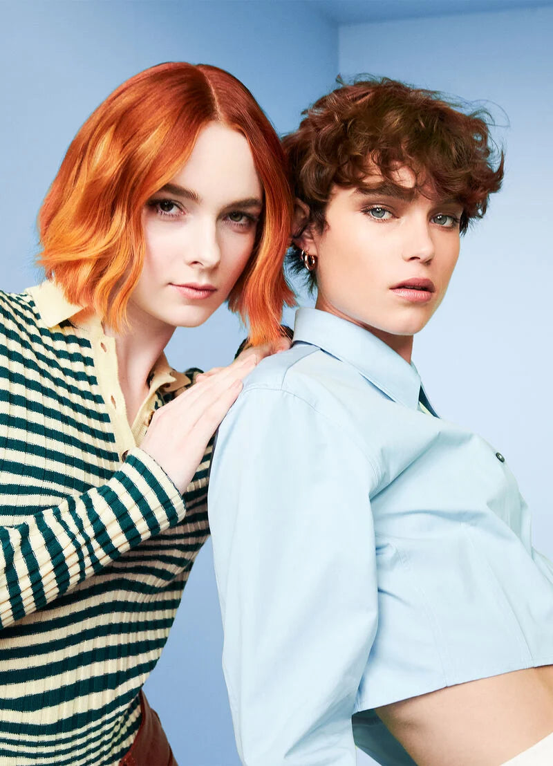 Two models posing in a studio setting against a light blue background. The model on the left has vibrant orange-red hair and wears a green and cream striped knit top over a yellow collared shirt.