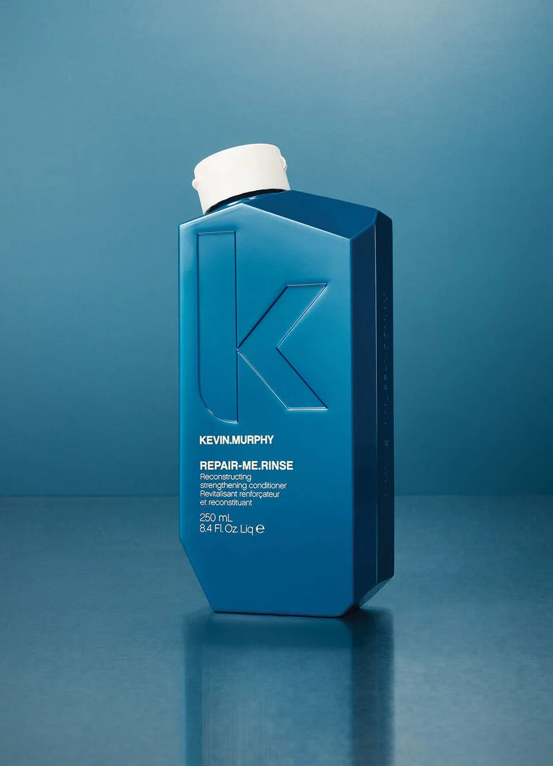Kevin Murphy Repair-Me Rinse bottle in a deep blue color with a white cap, standing on a reflective blue surface against a blue gradient background.