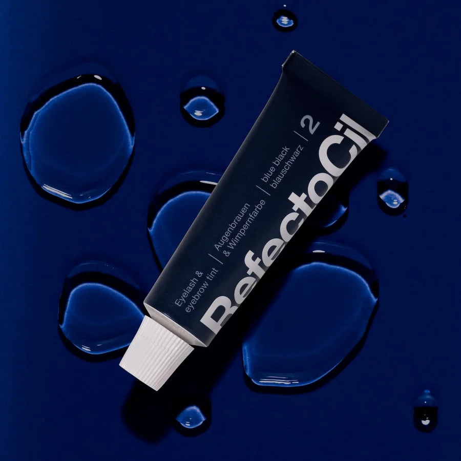 A tube of ReflectoOil 2 hair growth serum lies diagonally on a dark blue background decorated with glossy water droplets. The tube is black with white text and a white cap.