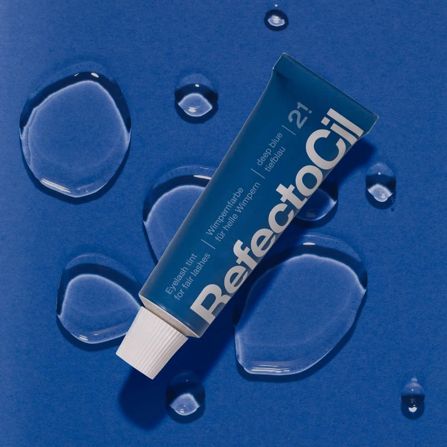A tube of RefectoCil 21 Deep Blue toothpaste lies diagonally on a textured blue background, surrounded by clear water droplets. The tube is dark blue with white text and a white cap.