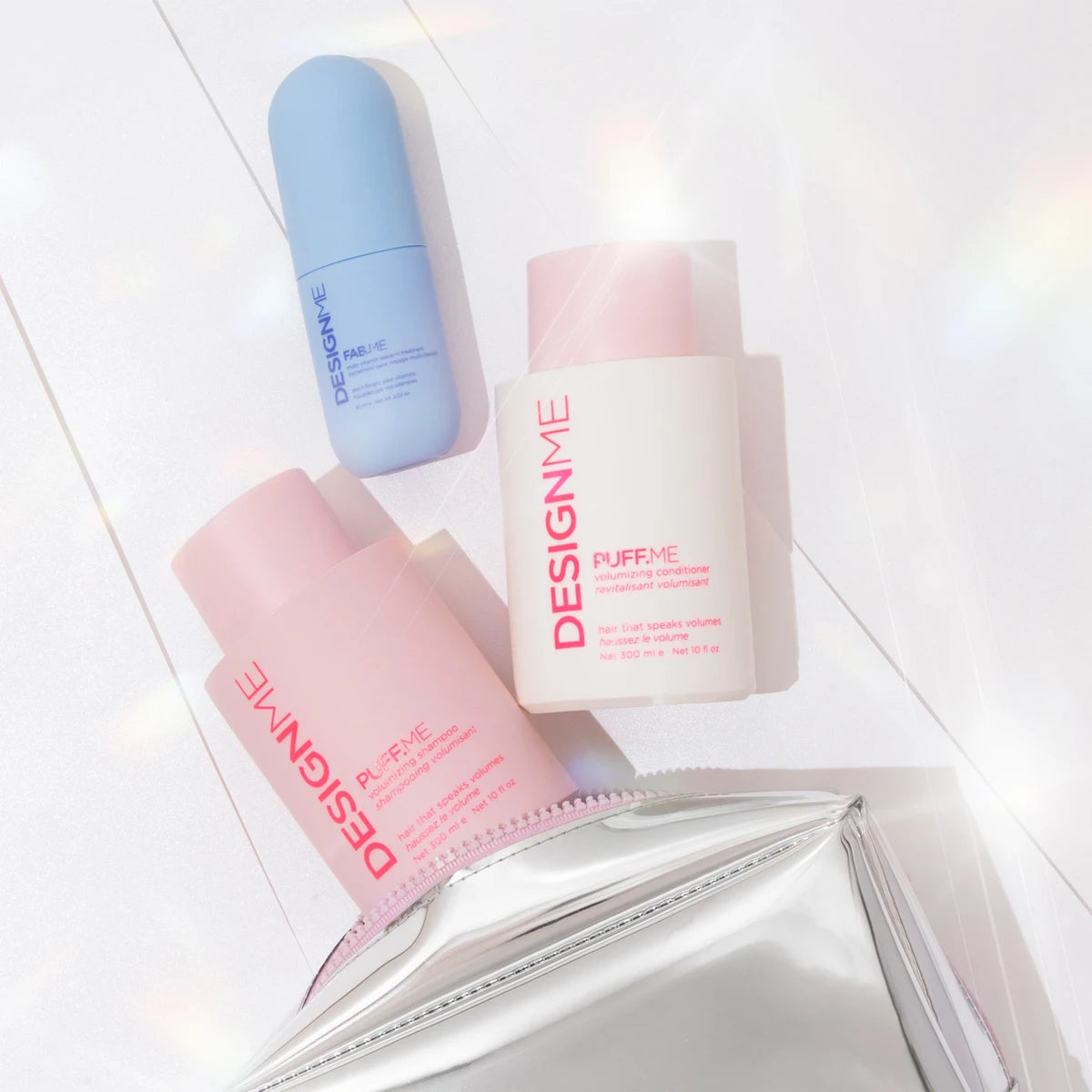 Three DESIGNME skincare bottles and a silver pouch arranged on a bright white surface with soft sunlight and lens flare effects. The set includes a light blue bottle, a white bottle with pink cap and pink text, a pink bottle with pink cap and pink text, and a reflective silver cosmetic pouch.