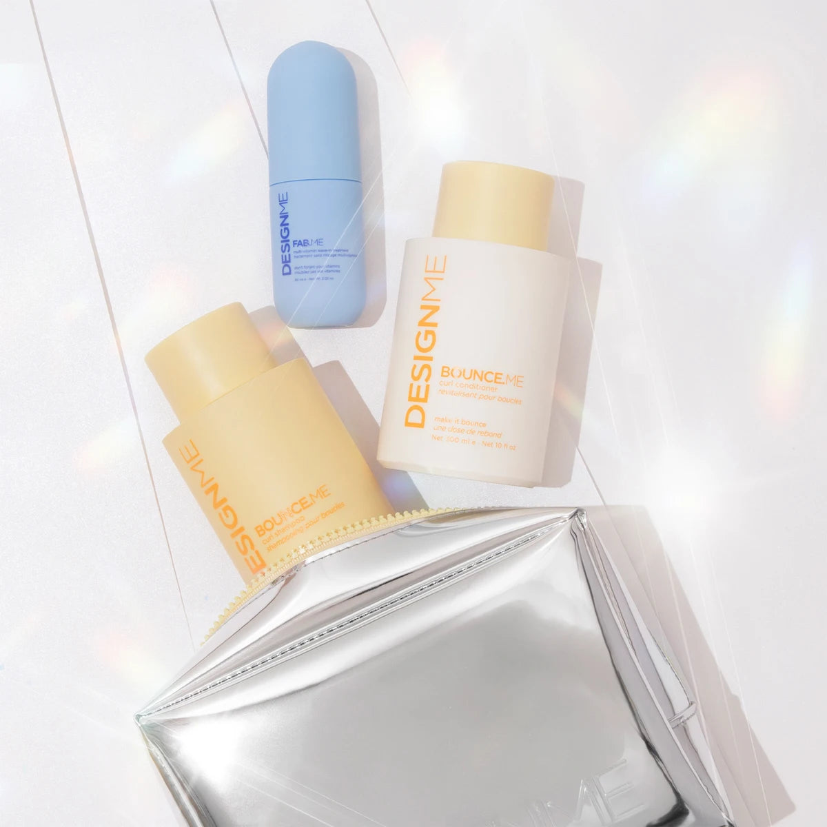 Three cosmetic bottles from the brand DESIGNME arranged on a white surface with a shiny silver pouch. The set includes a light blue bottle, a pale yellow bottle labeled Bouclée, and a cream-colored bottle labeled Blanche.