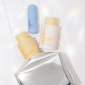 Three cosmetic bottles from the brand DESIGNME arranged on a white surface with a shiny silver pouch. The set includes a light blue bottle, a pale yellow bottle labeled Bouclée, and a cream-colored bottle labeled Blanche.