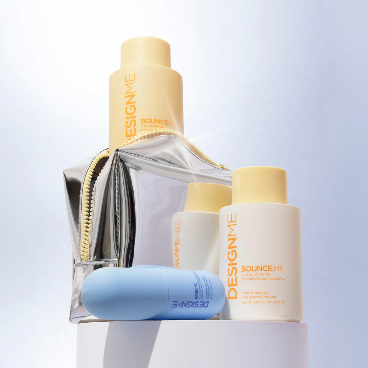 A set of DesignMe skincare products arranged on a white pedestal, including a large beige bottle, a smaller white bottle, a light blue tube, and a clear zip pouch with a metallic silver body and gold zipper.