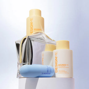A set of DesignMe skincare products arranged on a white pedestal, including a large beige bottle, a smaller white bottle, a light blue tube, and a clear zip pouch with a metallic silver body and gold zipper.