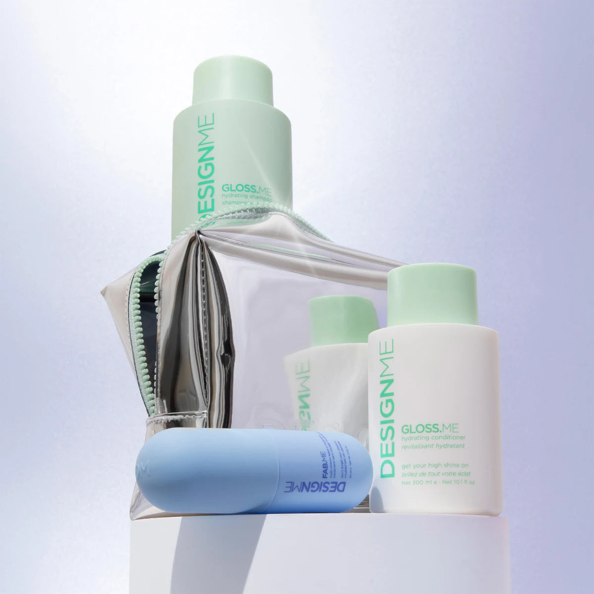 A set of DesignMe grooming products arranged on a white pedestal against a soft light purple background. The set includes a tall mint green bottle, a white bottle with a mint cap, a smaller white bottle, a light blue tube, and a clear zippered pouch with mint green trim.