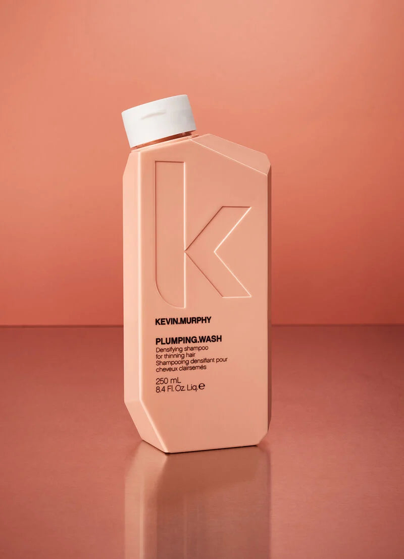 Kevin Murphy Plumping Wash shampoo bottle in a peach-pink color with a white cap, standing on a matching peach-pink reflective surface against a solid peach-pink background.