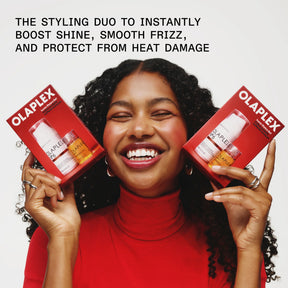 A smiling woman with curly hair holds two red Olaplex hair protection boxes in both hands against a light gray background, with promotional text at the top.