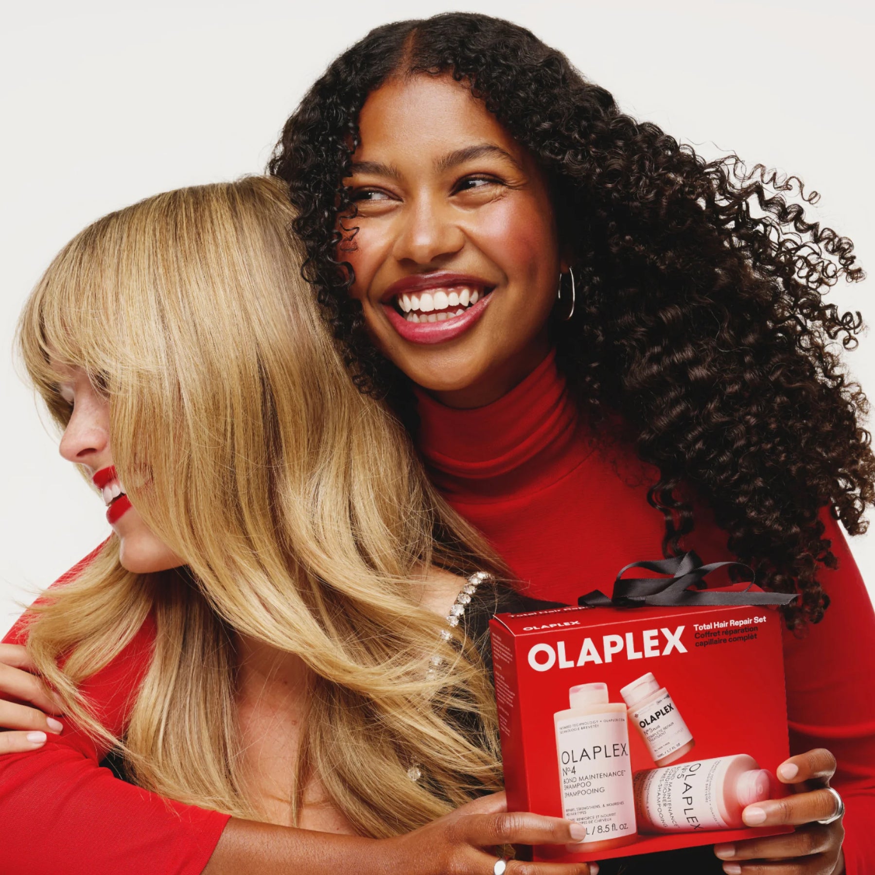 Two women hugging while holding an Olaplex hair building treatment box. The woman on the right has dark curly hair and wears a red turtleneck, while the woman on the left has blonde hair and a red top.