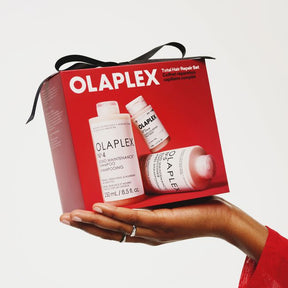 A hand holds an Olaplex Total Hair Repair Set gift box with a black ribbon. The red box features images of the included hair care products, including Olaplex K4 End Maintenance Shampoo and Olaplex Hair Building Treatment, against a light gray background.