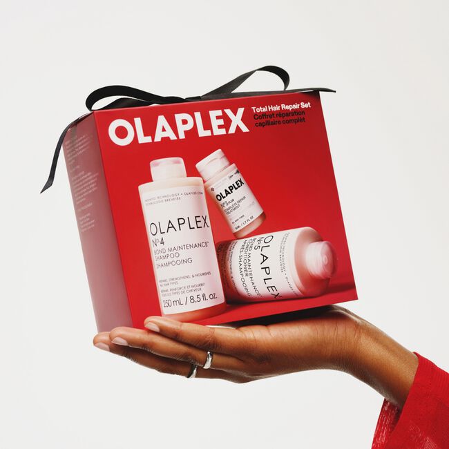 A hand holds an Olaplex Total Hair Repair Set gift box with a black ribbon. The red box features images of the included hair care products, including Olaplex K4 End Maintenance Shampoo and Olaplex Hair Building Treatment, against a light gray background.