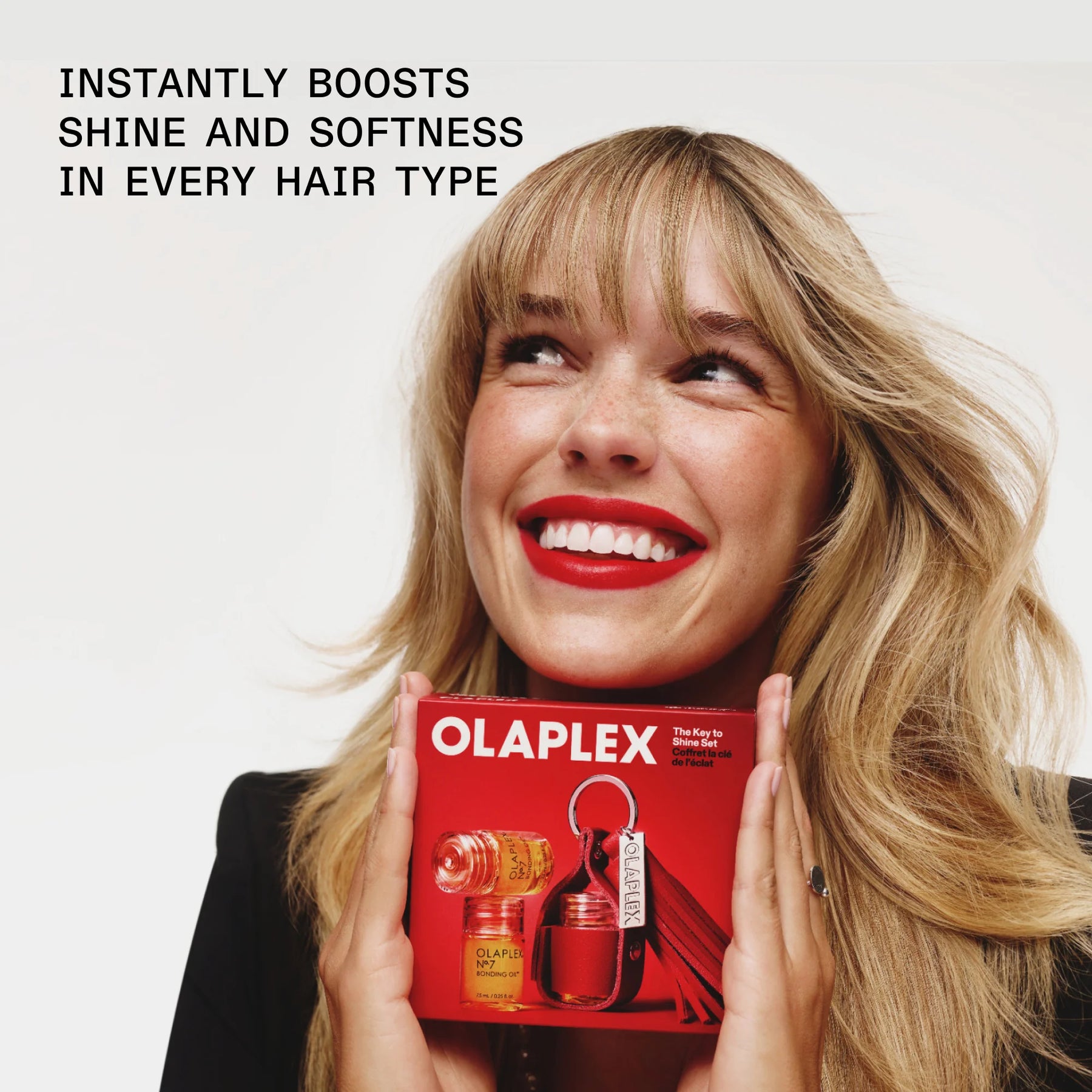 A smiling blonde woman holds an Olaplex box in front of her face against a light gray background, with promotional text at the top reading 'INSTANTLY BOOSTS SHINE AND SOFTNESS IN EVERY HAIR TYPE.'.