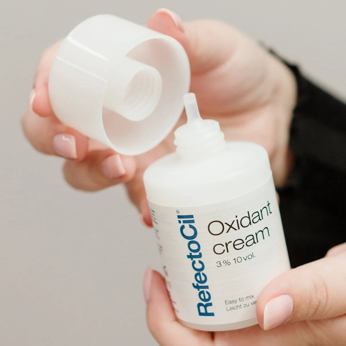 A close-up of a person holding a white bottle of RefectoCil Oxidant cream with its cap off, showing the applicator tip against a soft neutral background.
