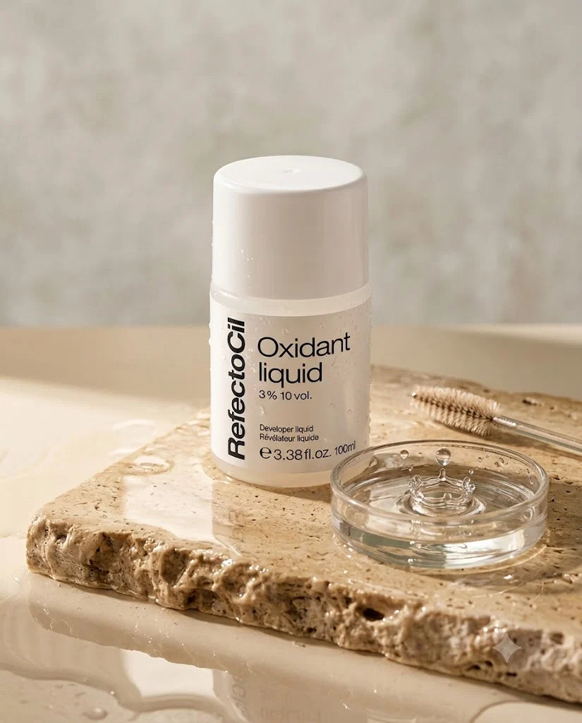 A bottle of ReflectoOil Oxidant liquid 3% 10 vol. developer liquid stands on a stone slab next to a clear glass lid and a makeup brush, set against a neutral beige background.
