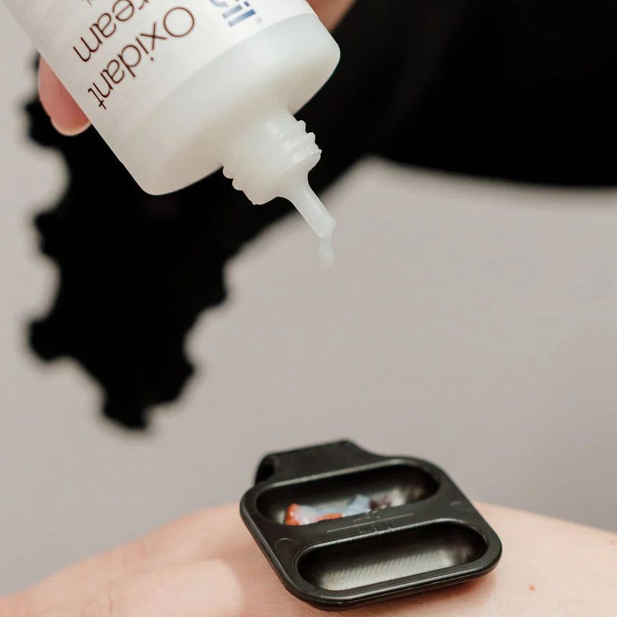 A close-up of a hand holding a white bottle labeled 'Oxidant cream' above a black dual-compartment spatula, with a single drop of white cream dripping from the tip onto skin.