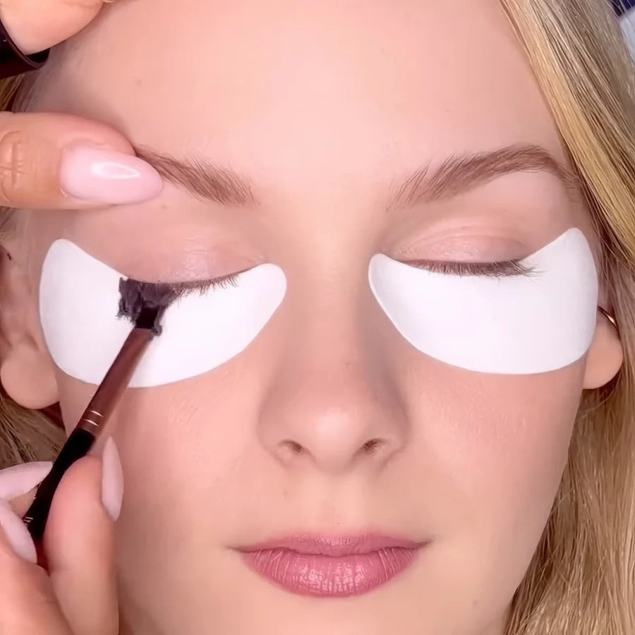 Close-up of a person applying dark eyeliner with a brush while wearing white under-eye patches.