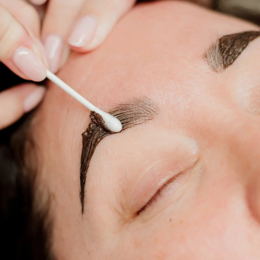 Close-up of a person's closed eye and eyebrow while a hand uses a cotton swab to clean or apply product to the eyebrow area.