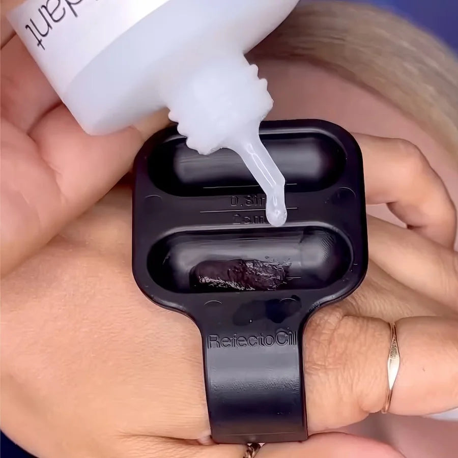 A close-up of a hand holding a black silicone ring mold while applying clear liquid from a white squeeze bottle into the mold. The mold has two oval cavities, one of which already contains a dark brown gel or wax.