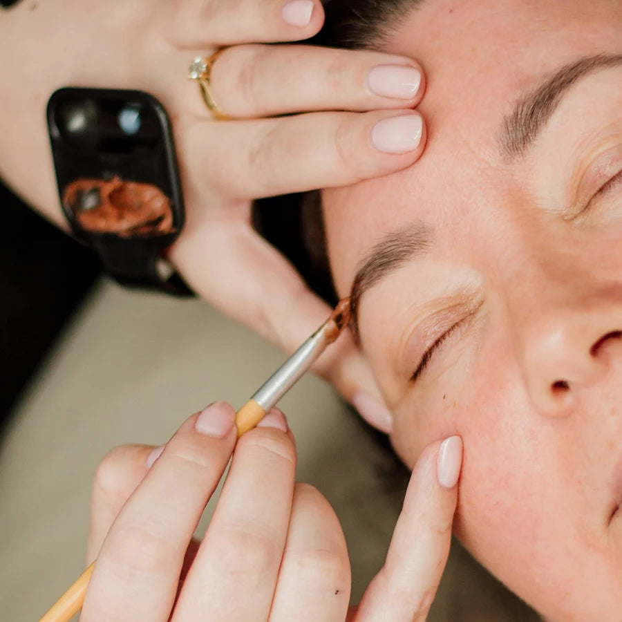 Close-up of a beauty practitioner applying brow makeup with a small brush to a client's eyebrow, showing detailed hand and face in a spa or salon setting.