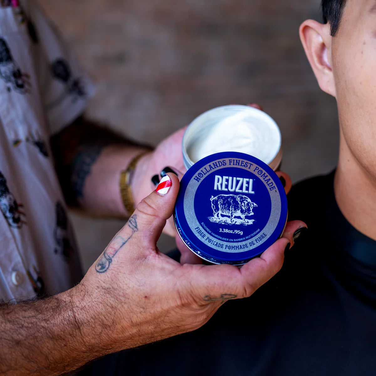 Person holding a Reuzel pomade container with another person partially visible in the background.