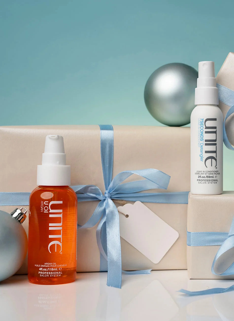 Two Unine hair care products, an orange spray bottle and a white mist bottle, styled on a white surface with beige gift boxes, light blue ribbons, and silver ornaments against a teal background.